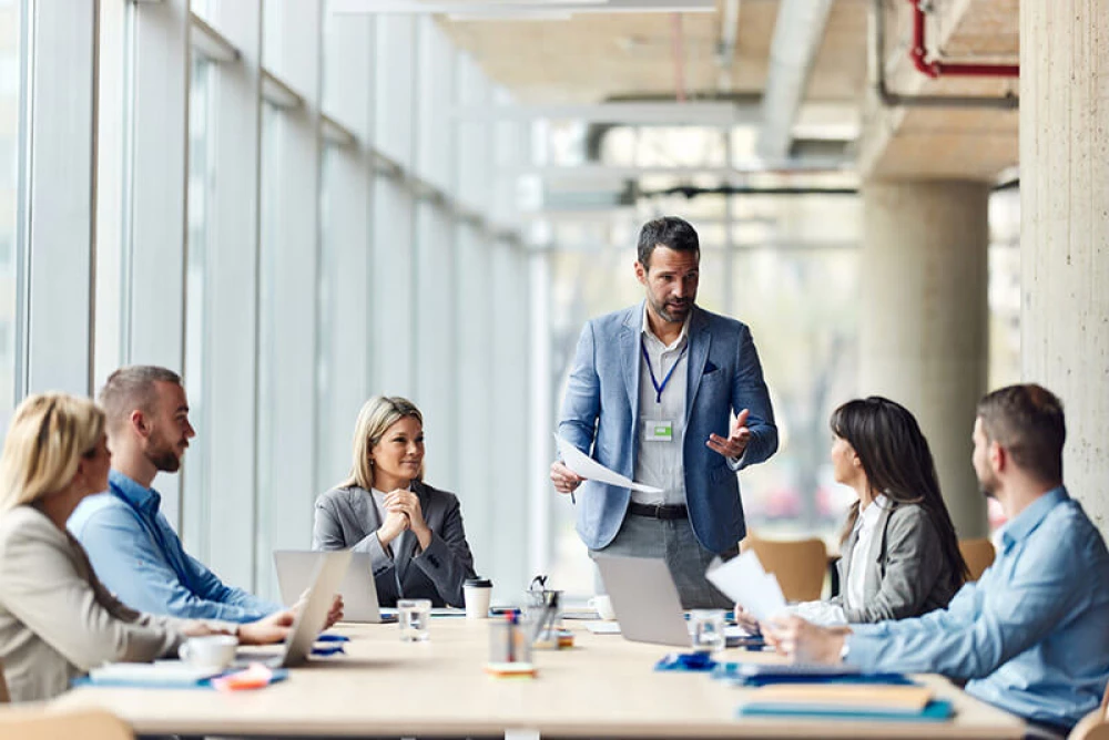 Man standing at a conference table with people sitting around it in suits