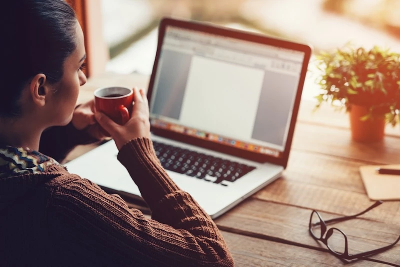 Woman holding coffee mug and looking at laptop.