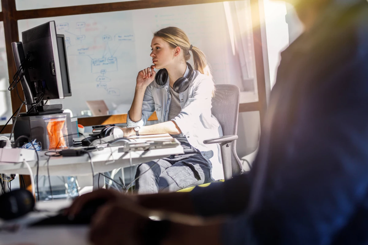Woman sitting at desk looking at computer monitor with headphones around her neck.
